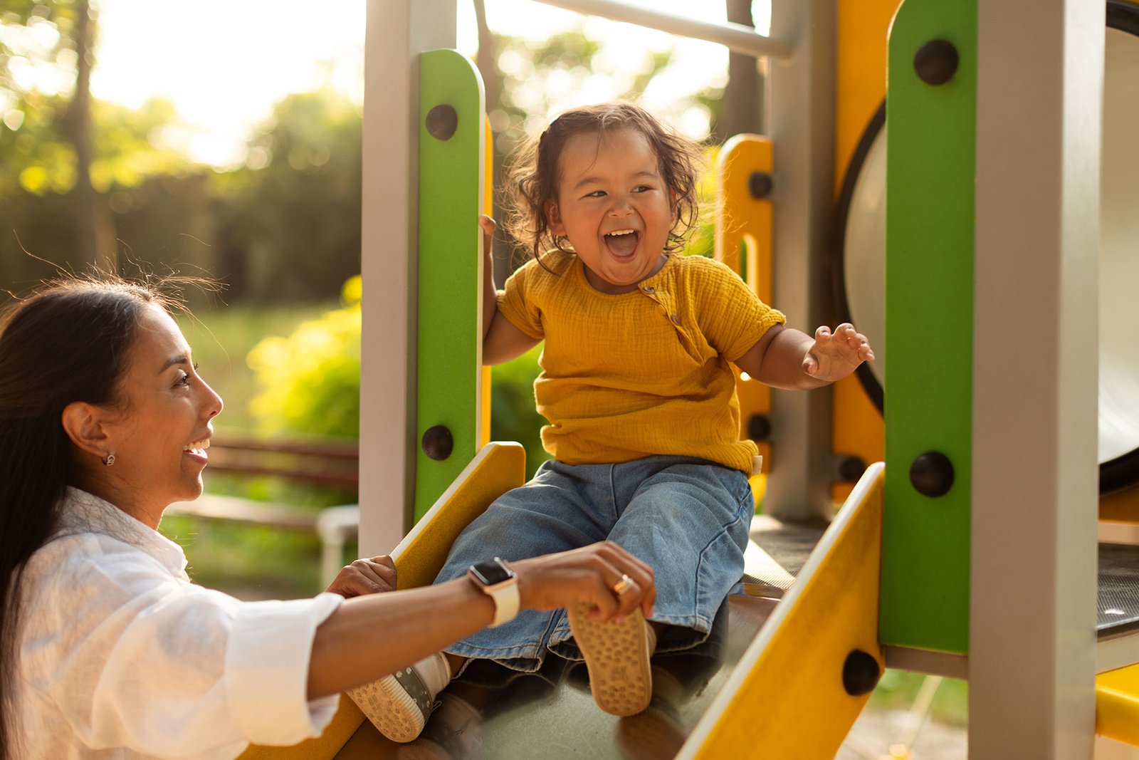 A mother laughing with her toddler on a playground slide
